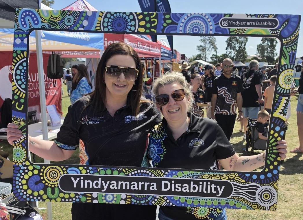 Two Yindyamarra Disability team members holding a painted Yindyamarra Disability photo frame at a community event