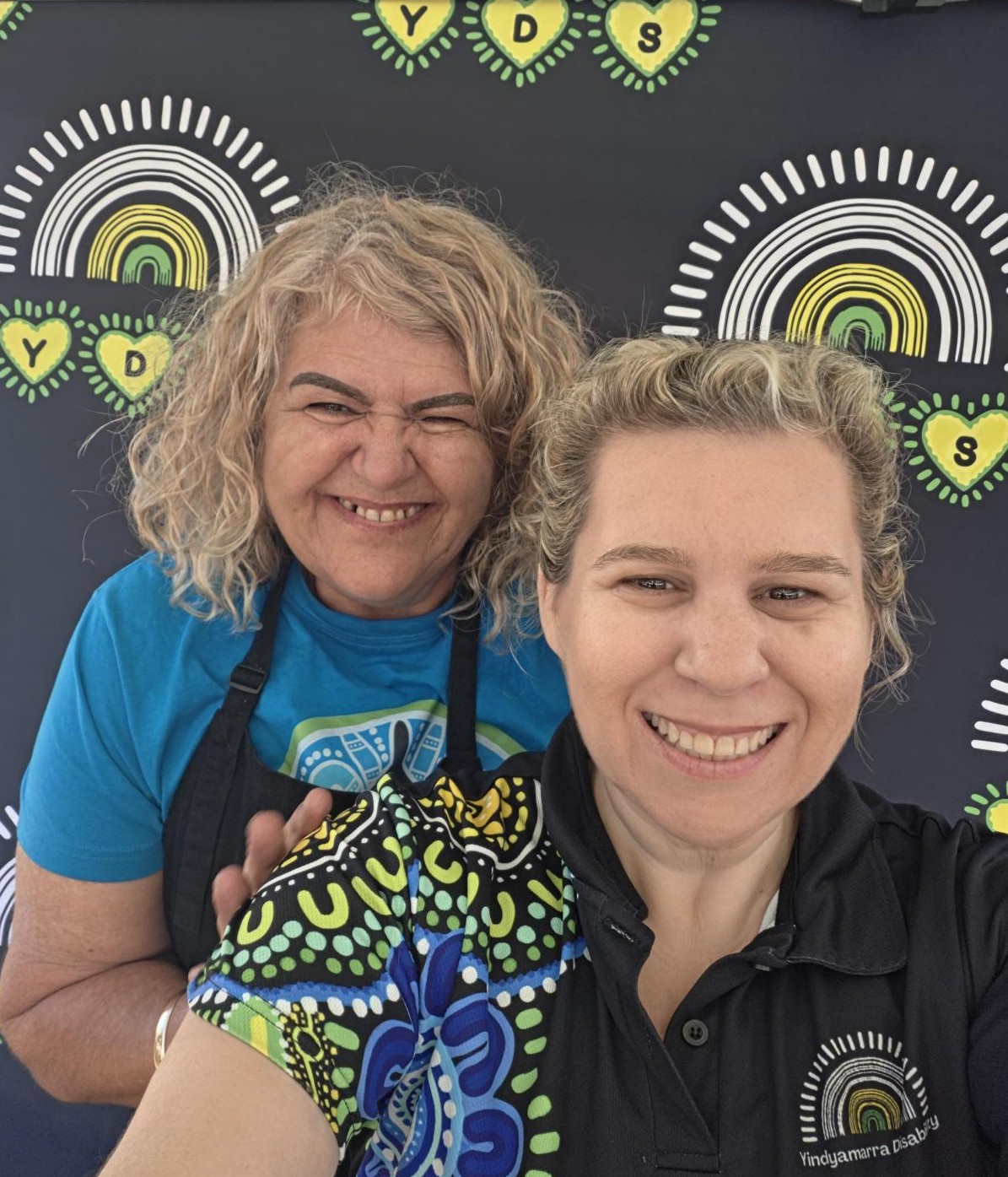 Two women laughing together in front of the Yindyamarra Disability backdrop